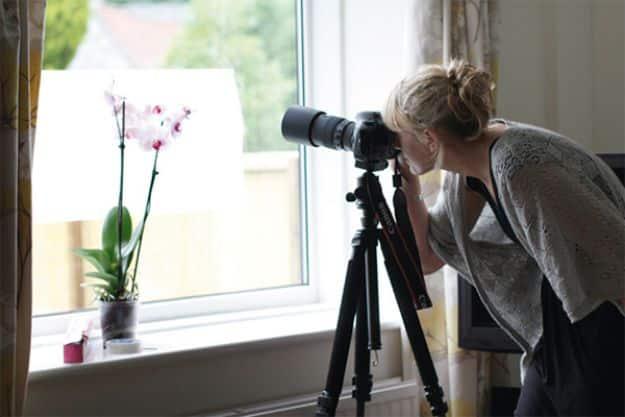Window lightbox, a woman at a window with a tripod mounted camera photographing a pale orchid lit from behind