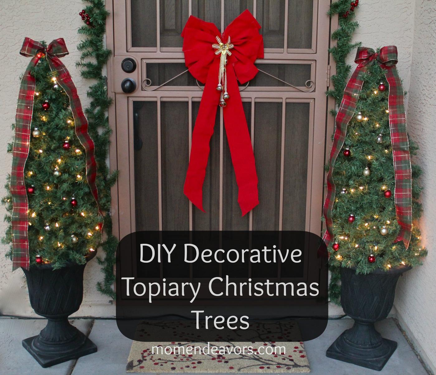 Two lit topiary Christmas trees with plaid ribbon and ornaments in black urn planters flanking a front door with a red bow