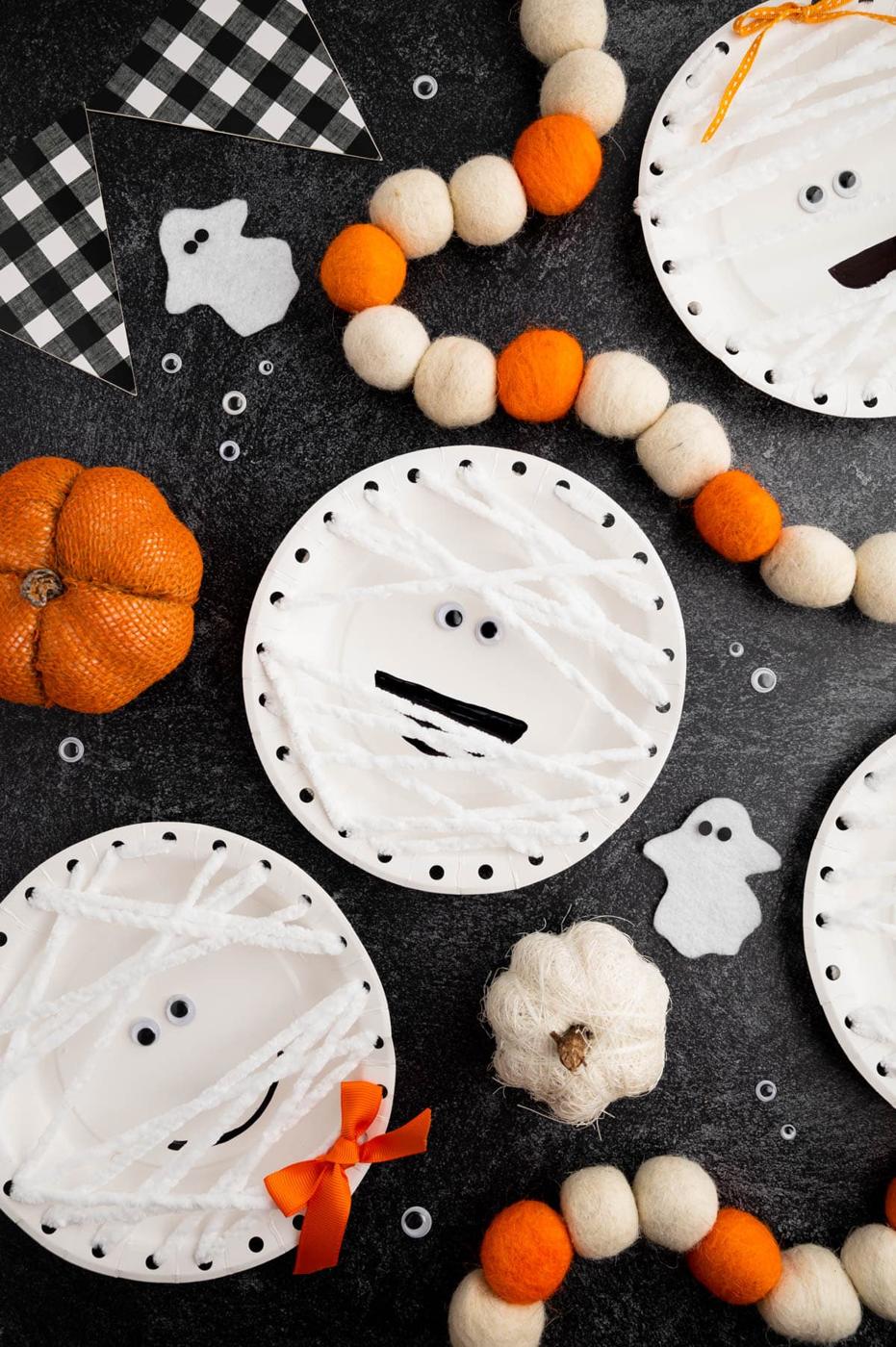 White paper-plate mummy crafts laced with white yarn, googly eyes and black mouths among orange Halloween decorations