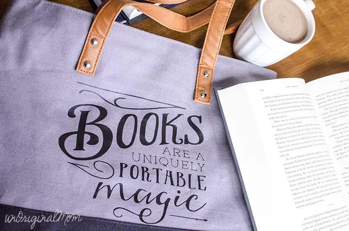 A gray canvas tote with leather handles lettered books are a uniquely portable magic in black beside an open paperback