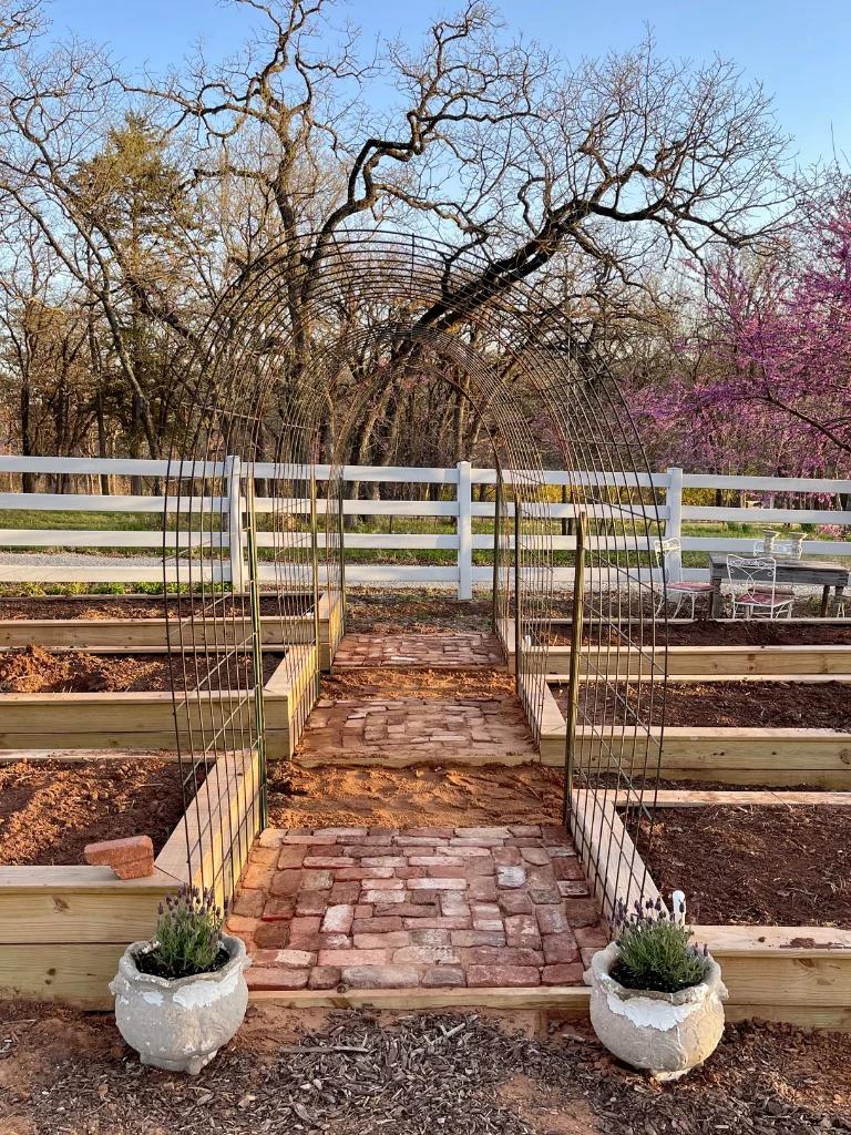 Red brick garden path bedded in sand, running between raised wooden vegetable beds toward an archway