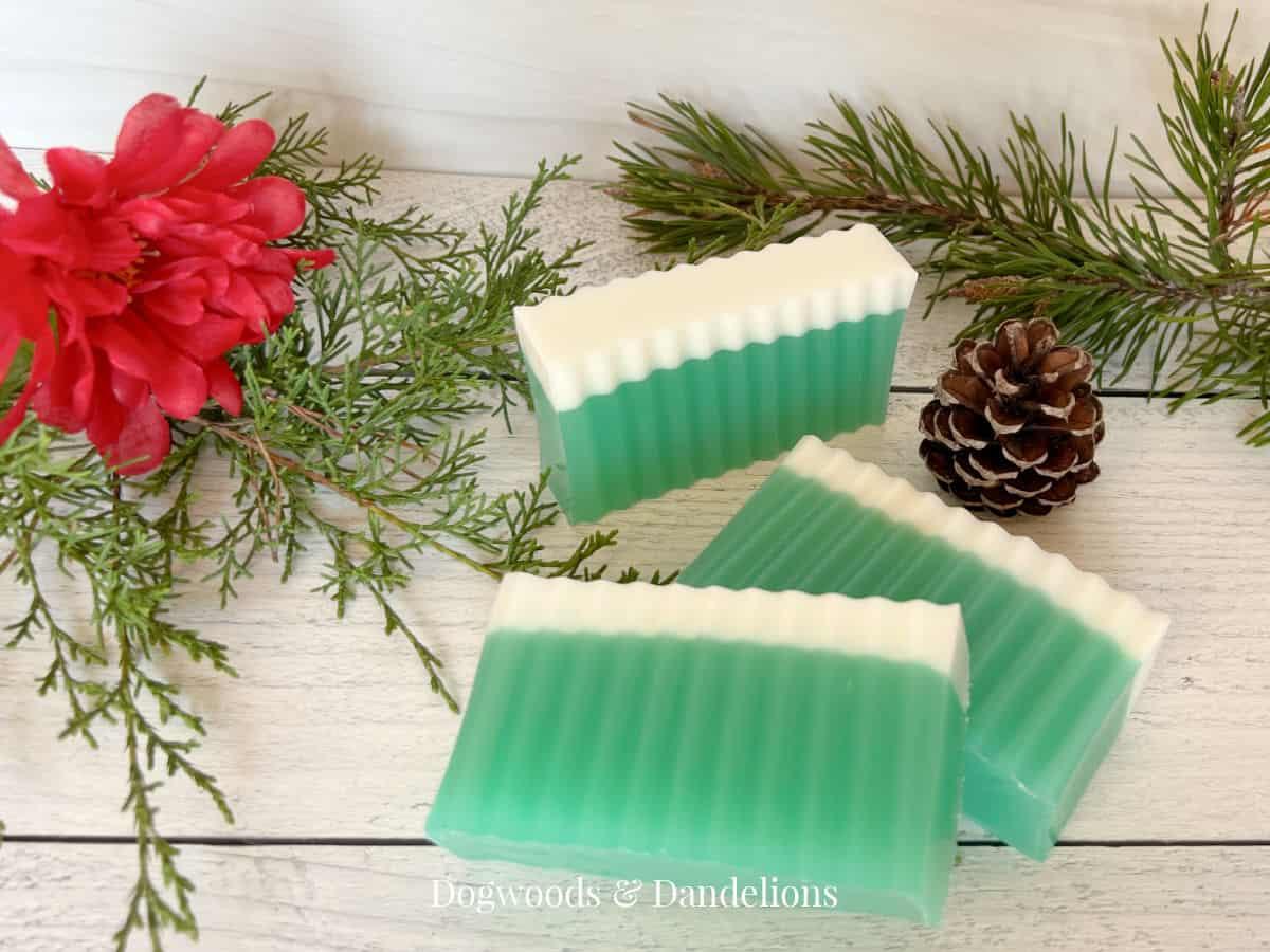 Three bars of green and white layered winter forest soap with crinkle cut edges beside a pinecone, evergreen and a red flower