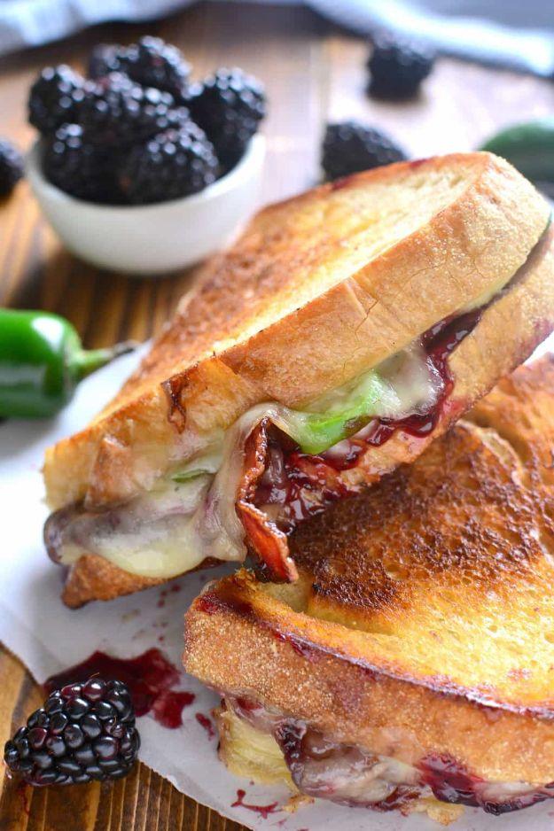 Blackberry bacon grilled cheese cut open on a plate showing melted cheese, bacon and dark jam, berries behind