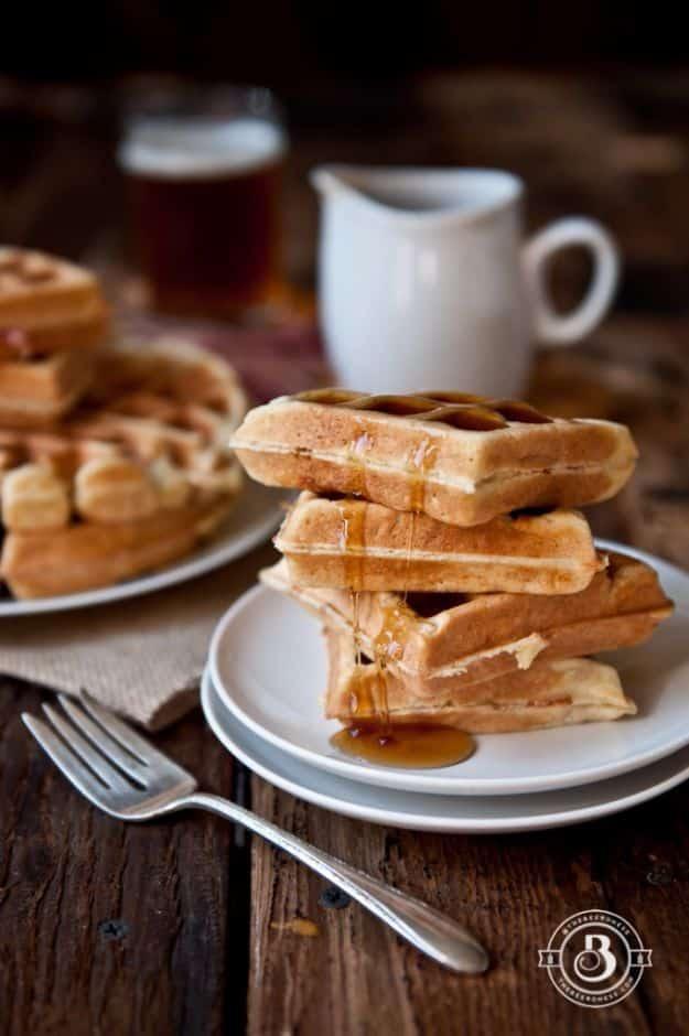 Maple bacon beer waffles stacked on a white plate with syrup, a glass of beer and a jug of syrup behind