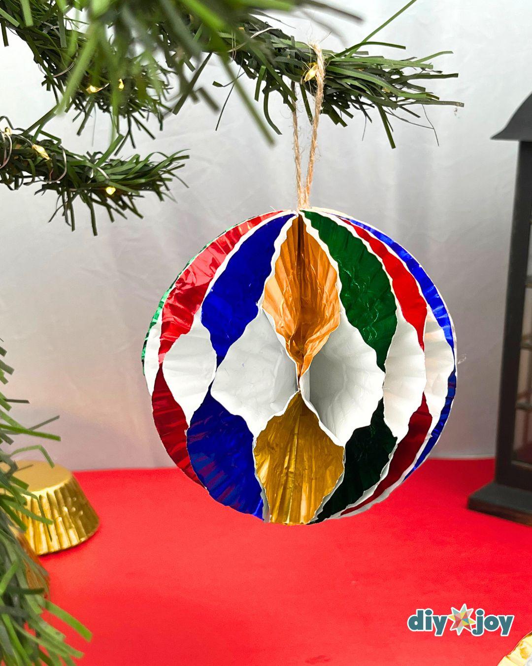 Christmas Tree Decorations - a red, blue, green, white, and gold striped ornament hanging from a pine branch with fairy lights