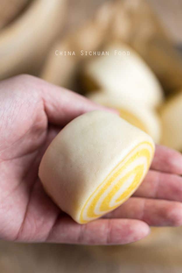 A hand holding a raw swirled pumpkin bun, showing the yellow and white spiral inside it before steaming