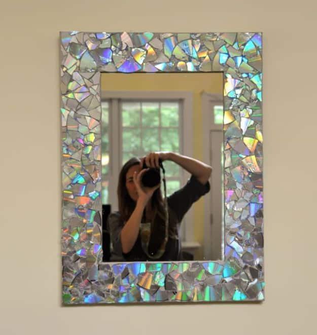 Rectangular wall mirror with its frame covered in broken CD shards, reflecting the room and a doorway behind