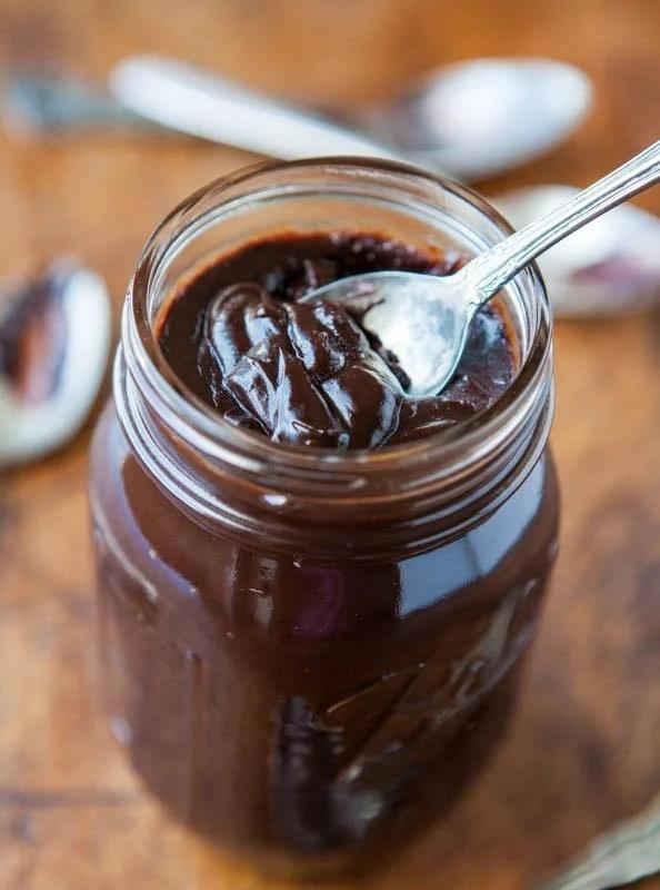 Glass jar of thick dark homemade hot fudge sauce with a silver spoon lifting a glossy ribbon of it out of the jar