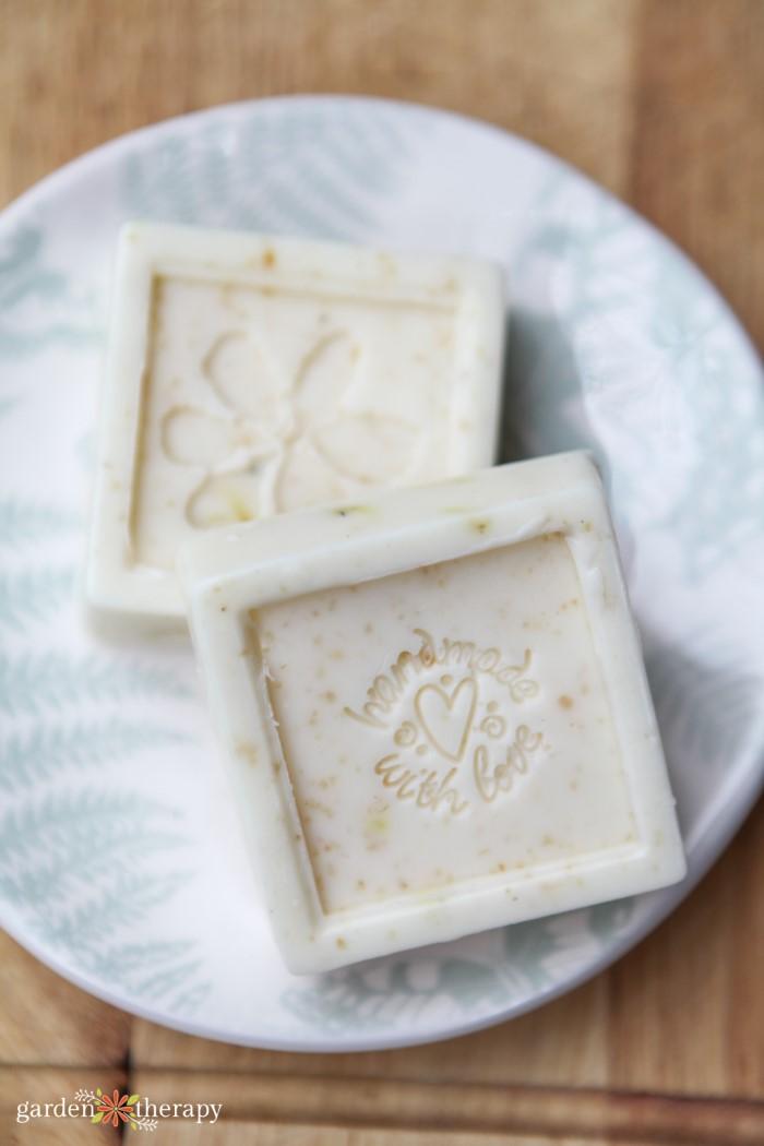 Two square cream colored soap bars embossed with a flower and a handmade with love stamp on a fern printed plate