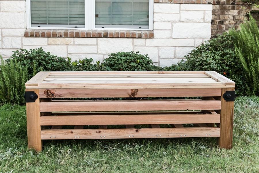 Outdoor storage ottoman built from cedar slats with black metal corner brackets, on grass beside a brick house wall
