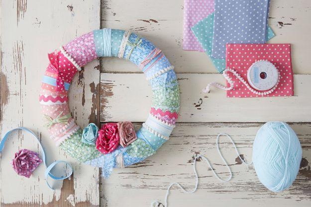 Fabric wreath wrapped in pastel strips, trims and rolled fabric roses, laid on a distressed white wood table