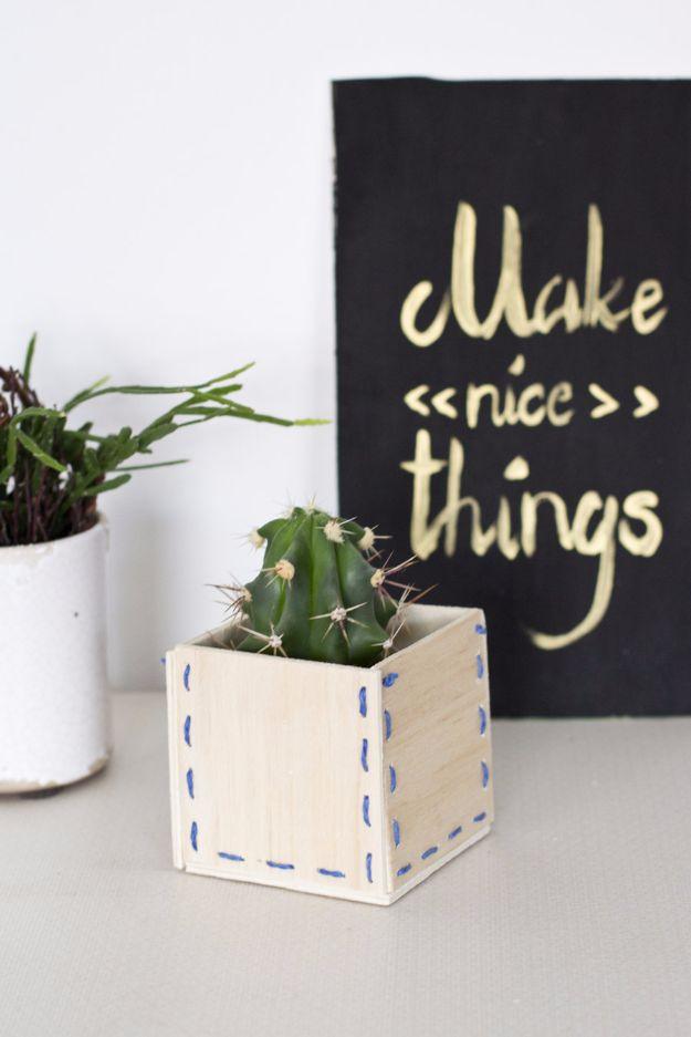 Small pale wooden box planter laced at the corners with blue yarn stitches, holding a round cactus on a desk