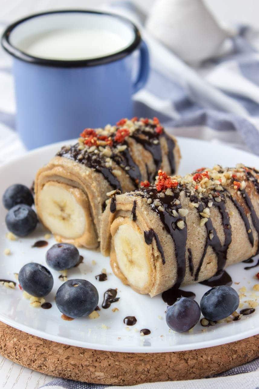 Peanut butter banana roll ups sliced on a plate with chocolate drizzle, blueberries and a cup of milk