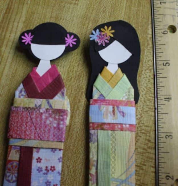Two paper kimono dolls with black paper hair and flower pins, in pink and green patterned kimono, on a wooden ruler