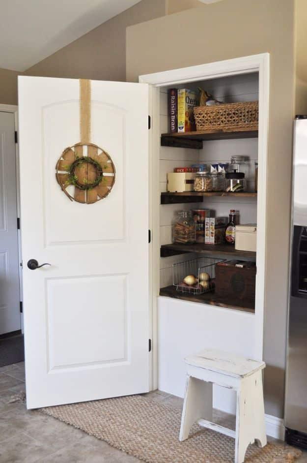 Open white pantry door with a burlap hung wreath, dark stained wood shelves, jars and baskets inside, white stool