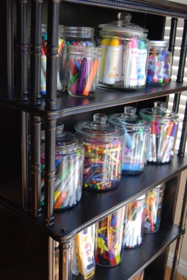 Jar storage for art materials, glass jars of markers crayons and paint pens lined along the shelves of a dark bookcase