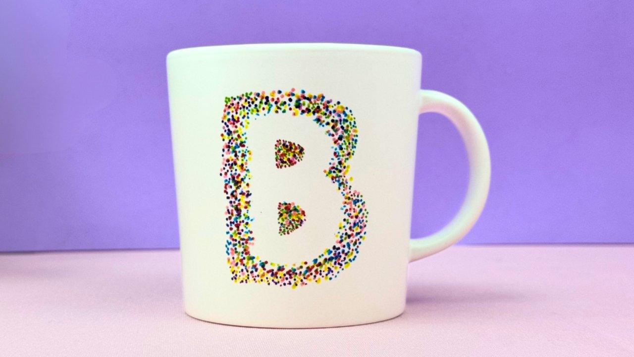 Sharpie mug craft, a white mug with a large letter B filled in with tiny multicolored marker dots on a lilac background