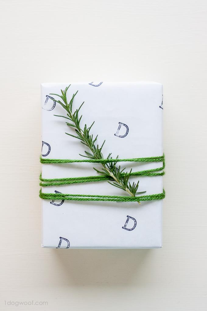 White wrapped box stamped with the letter D in diagonal rows, wound with green yarn and topped with a sprig of fresh rosemary