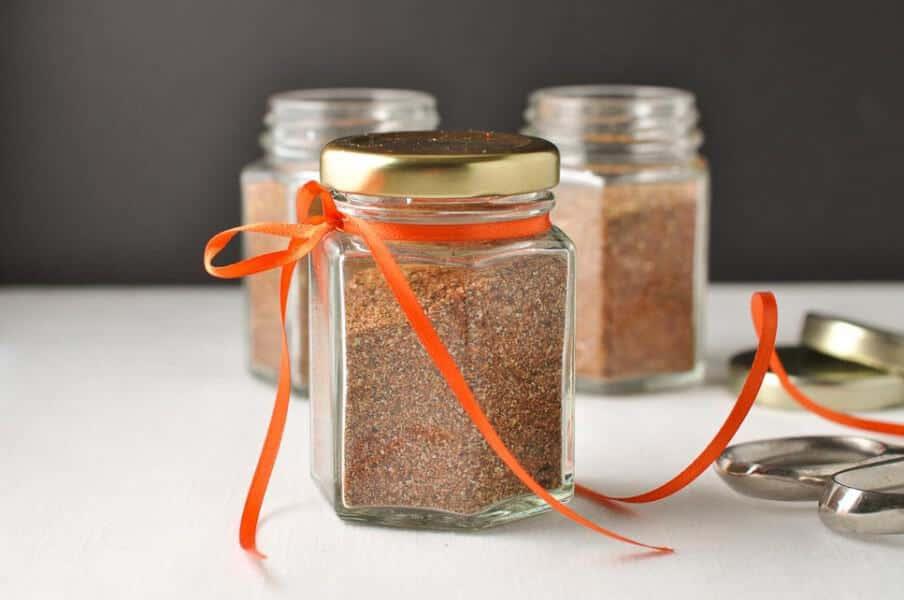 Small glass jar of reddish homemade taco seasoning mix with a spoonful tipped out onto a wooden board