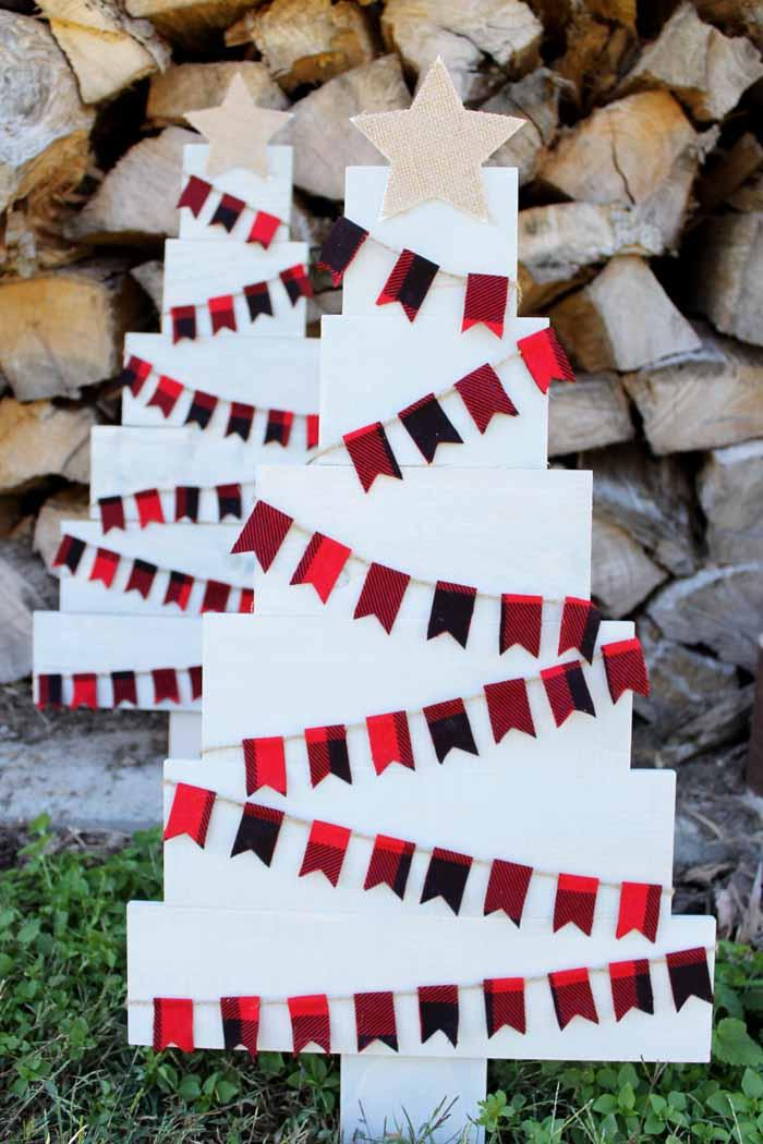 Two white washed pallet wood Christmas trees wrapped in red and black plaid pennant banners with burlap stars, by a wood pile