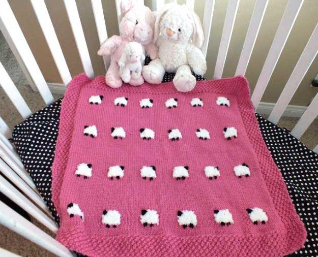 Pink baby blanket with rows of small fuzzy white sheep, hung over a crib rail with stuffed animals sitting on it
