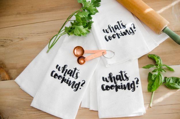 Three white flour sack tea towels lettered with what's cooking in black script, with copper spoons and herbs