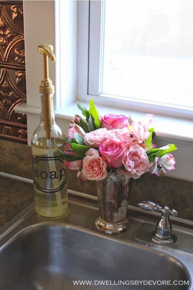 Glass syrup bottle used as a soap dispenser with a gold pump and a soap label beside a sink and pink roses
