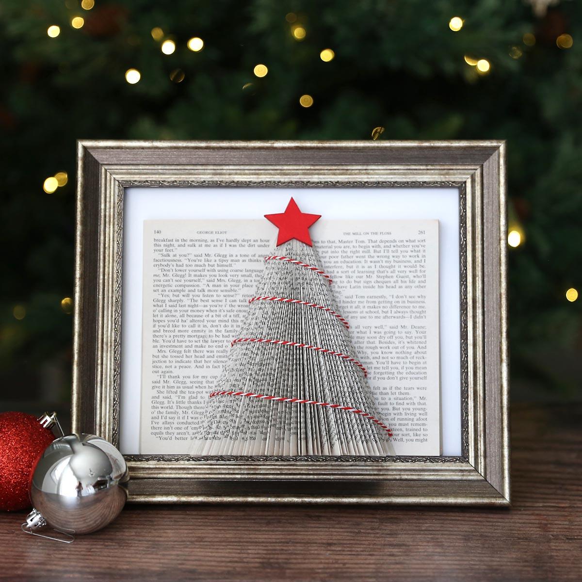 A folded book page Christmas tree with a red star and red and white twine garland in a silver frame in front of a lit tree