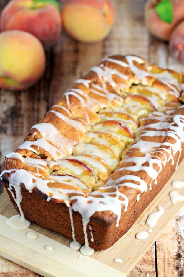 Pecan peach quick bread drizzled with white icing on a board, fresh whole peaches sitting behind the loaf