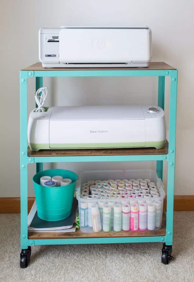 Turquoise metal rolling cart with wooden shelves holding a printer, a cutting machine, a bin of craft paint and vinyl rolls
