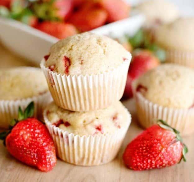 Keto strawberry muffins stacked in white paper liners with fresh berries and a white dish of them behind