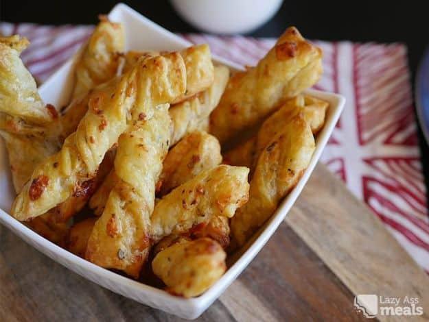 Super easy puff pastry cheese twists, golden twisted pastry sticks piled up in a white serving bowl.