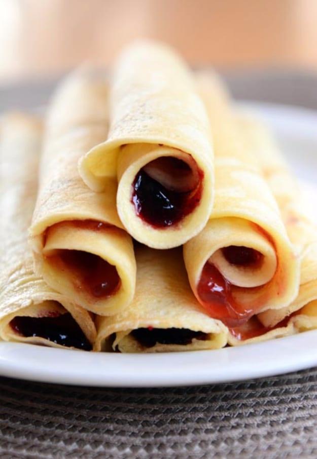 Rolled blender pancakes stacked on a white plate with red jam visible inside every rolled up pancake