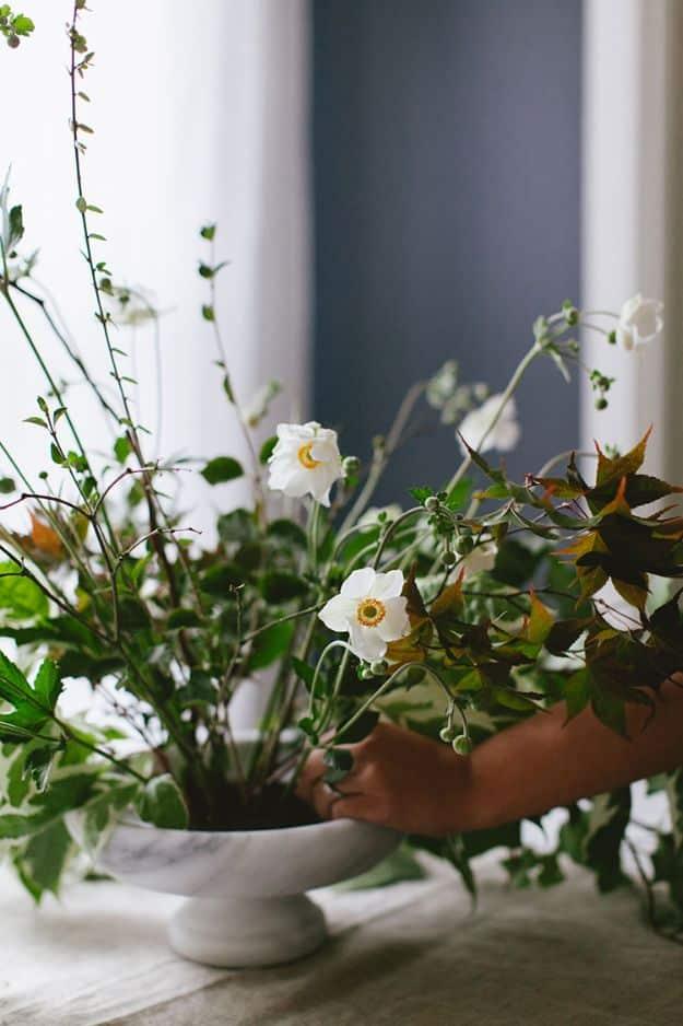 Shallow white footed bowl holding foraged branches and white anemones, with a hand adjusting a stem at the edge