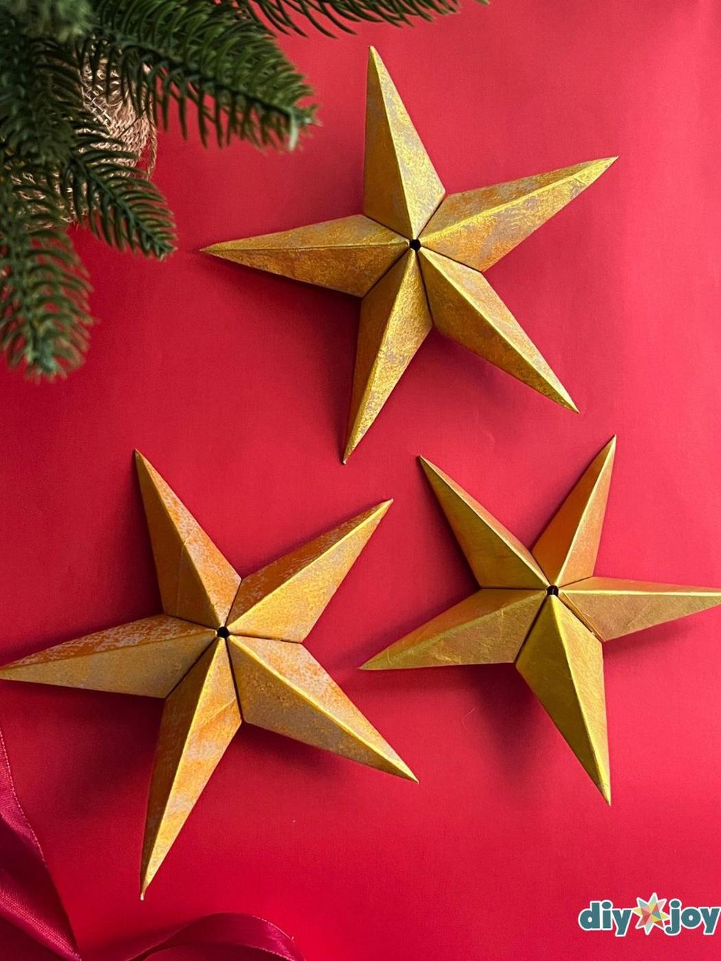 Three 3D five point star ornaments folded from a paper bag and painted textured metallic gold on a red background