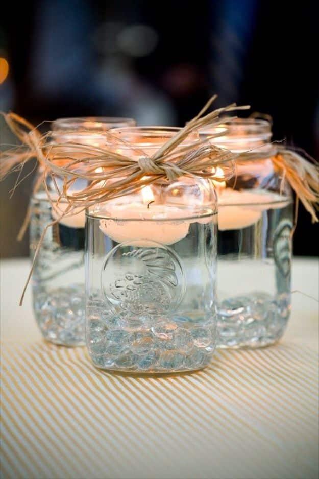 Group of mason jars filled with water and floating lit candles, tied with raffia bows over glass pebbles