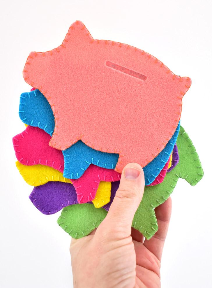 Hand holding a stack of blanket stitched felt piggy banks in coral, blue, pink, yellow, purple and green