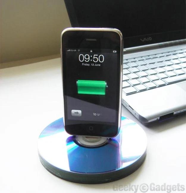 Phone dock made from a stack of glued CDs holding an iPhone upright and charging, on a desk beside a silver laptop
