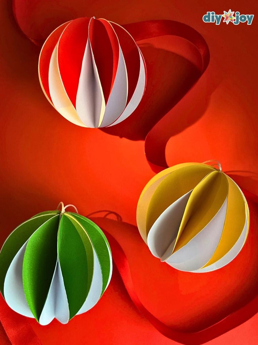 Three folded paper ball ornaments in red and white, green and white and yellow and white on a red background with ribbon