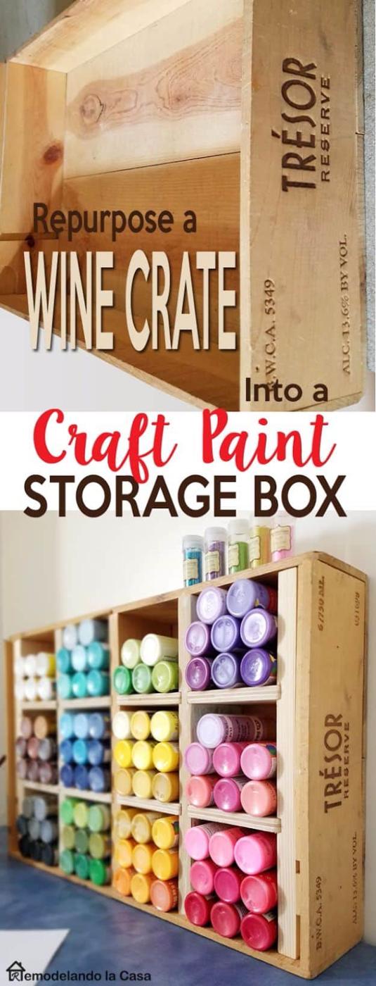 Wine crate on its side fitted with wooden shelves holding craft paint bottles lid out in rainbow order, glitter pots on top