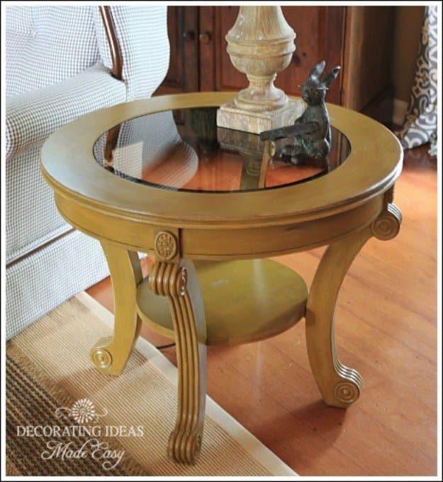 Round wooden center table painted gold with a glass top, holding a carved lamp base in front of a wood cabinet