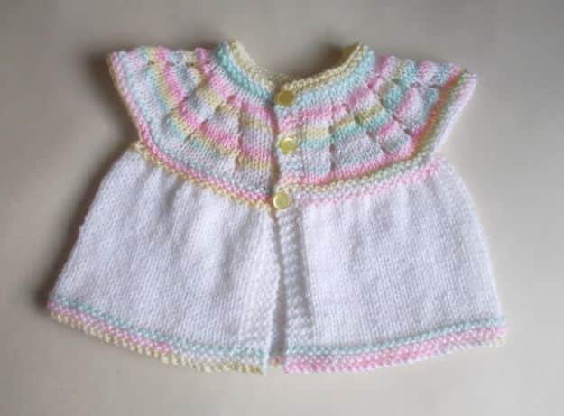 White knitted baby top with a pastel striped yoke and small buttons down the front, laid flat on a pale surface