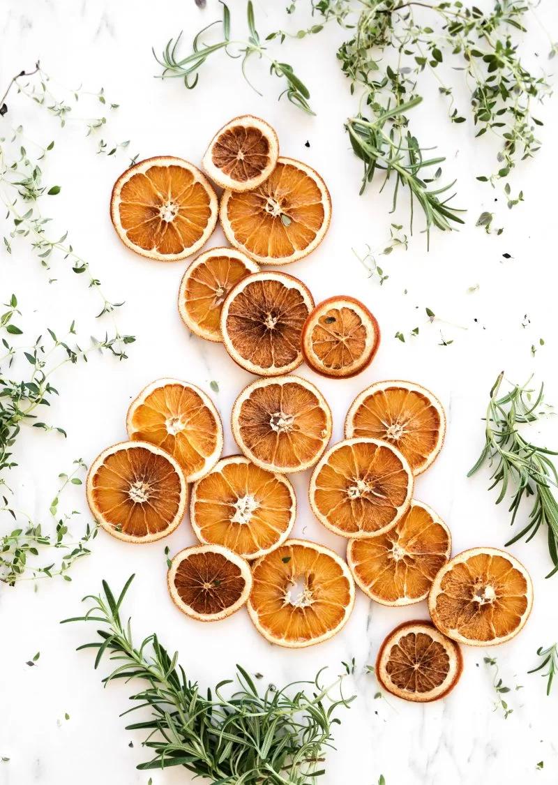 Dried orange slices in deep amber scattered with fresh rosemary and thyme sprigs on a plain white surface