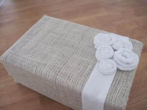 Shoe box wrapped in cream burlap with a wide ribbon band and three rolled fabric rosettes on the lid