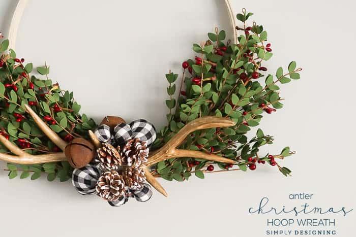 A wooden hoop wreath with faux antlers, a buffalo check bow, snowy pinecones and bells at the bottom and boxwood to each side
