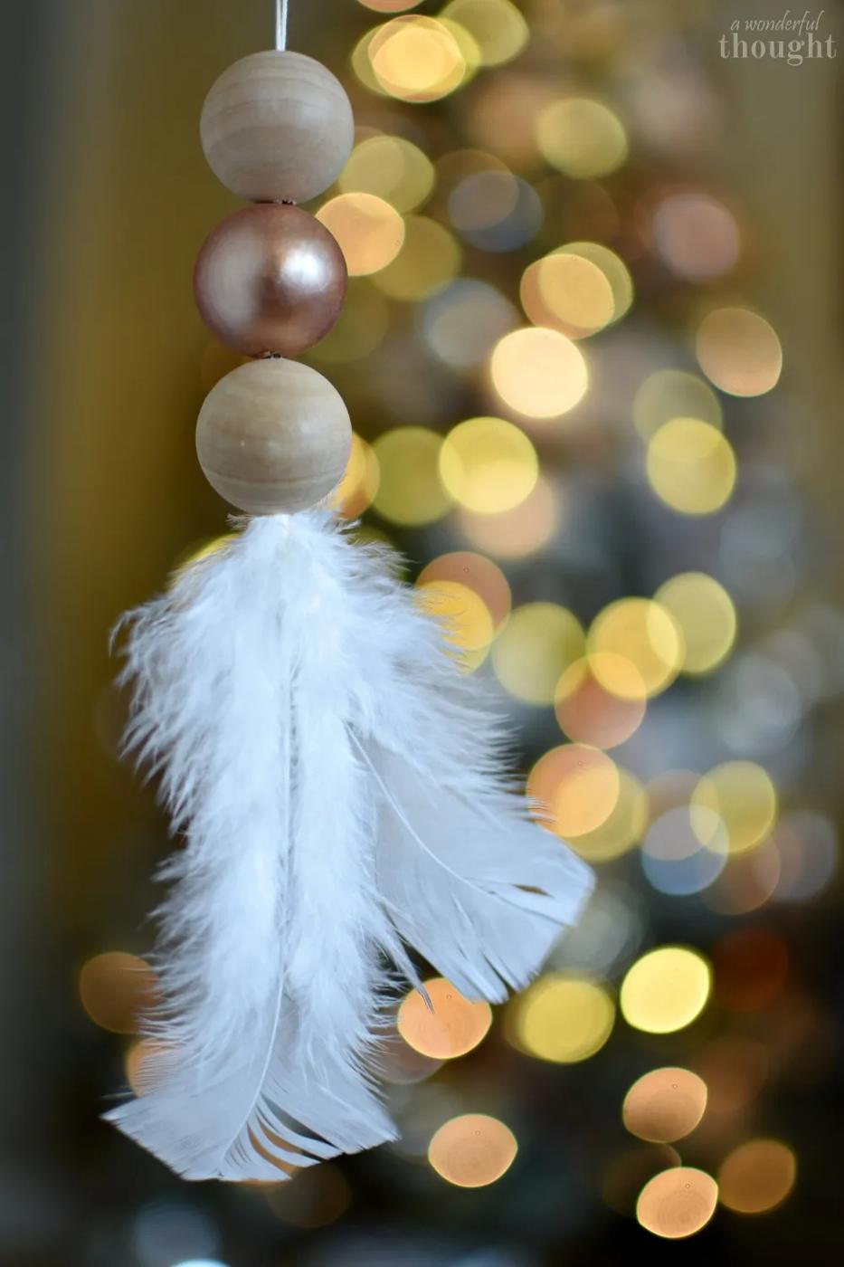 Two natural wood beads and one copper painted bead strung above a white feather, hanging in front of blurred Christmas lights