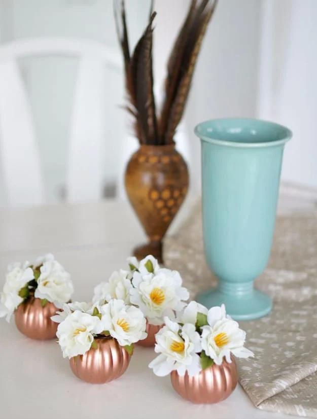 Four small copper painted pumpkin pails holding white silk flowers on a table beside a pale aqua vase