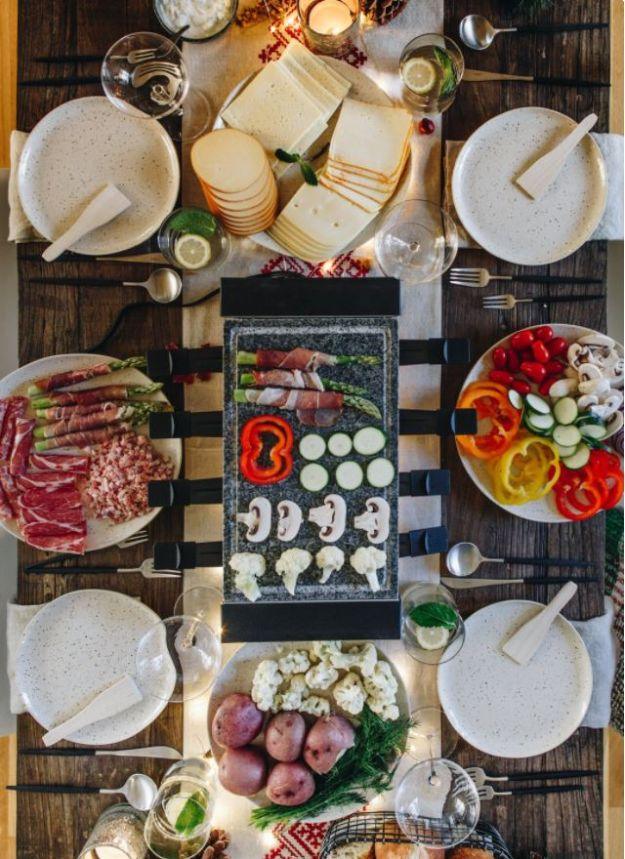 Overhead raclette spread with cheeses, cured meats, boiled potatoes, tomatoes and small plates on a striped runner