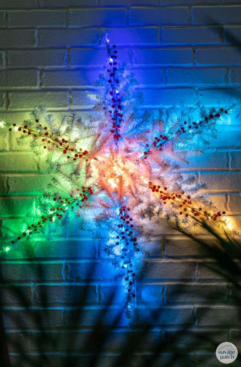 Large six point snowflake made from white pine branches wrapped in string lights glowing on a dark exterior wall