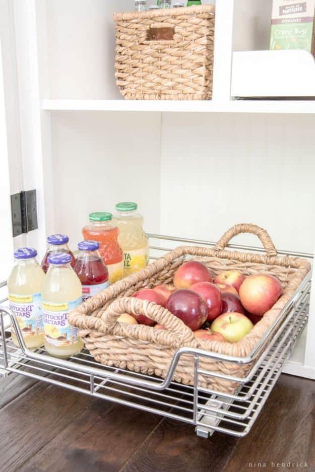 Chrome pull out wire basket drawer in a white pantry holding a woven tray of apples and glass juice bottles